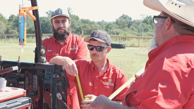 Two crew members in red company shirts at the open tailgate of a work truck, a caulk gun in a rack and a folding rule in hand.