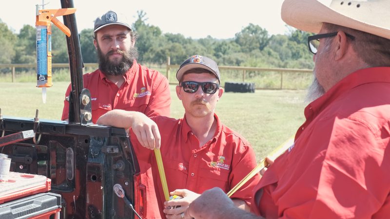 Bell crew in red company shirts, caps and sunglasses gathered at the open tailgate of a work truck loaded with toolboxes and a bucket