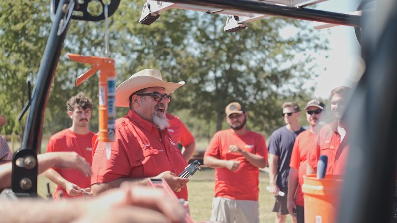 Lance Bell, in a straw hat, glasses and a red company shirt, talking with the crew outdoors beside a work truck with a ladder rack