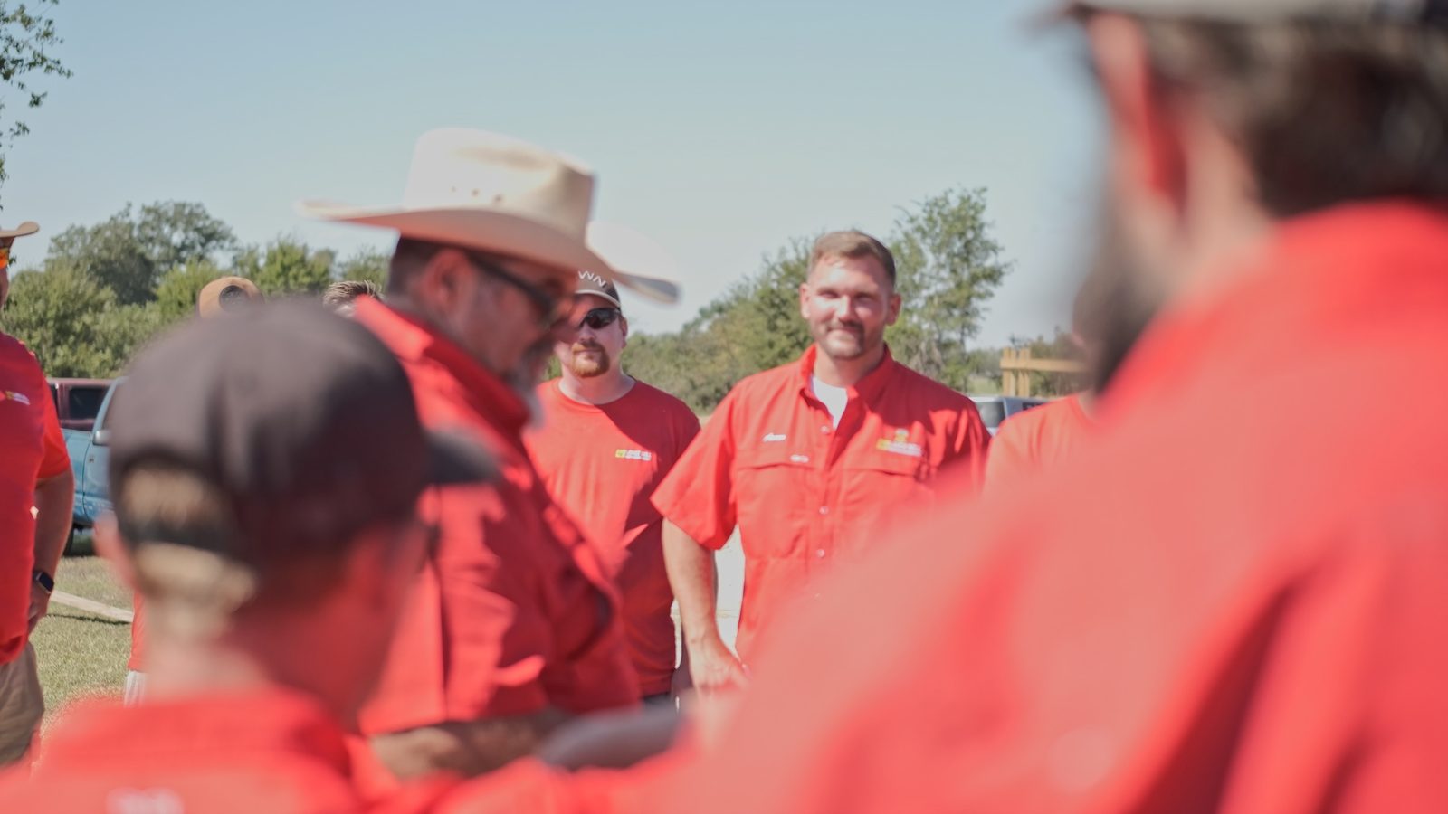 Bell crew members in red company shirts standing outdoors in the sun, one of them smiling toward the camera.
