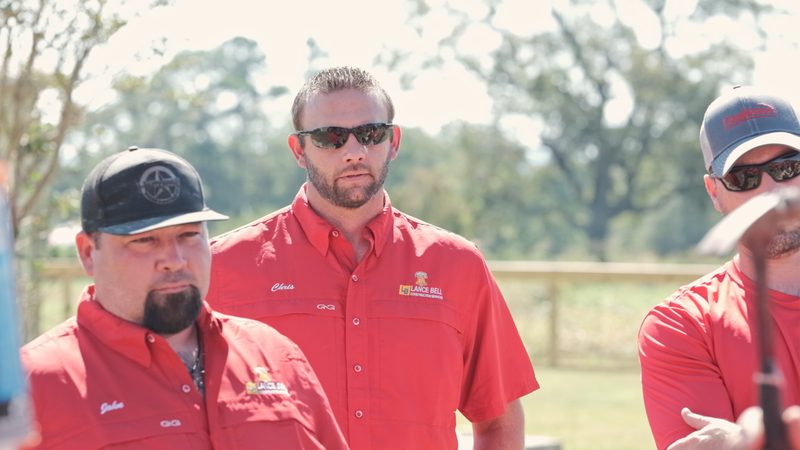 Bell crew members in red company shirts outdoors, one in a dark cap and one in sunglasses, an open field and a fence line behind