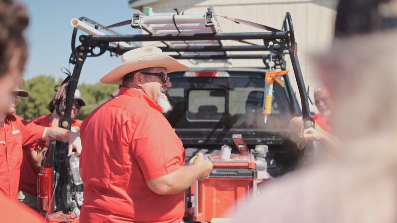 A Bell crew member in a straw hat at the open tailgate of a work truck fitted with a ladder rack, tool cases on the tailgate