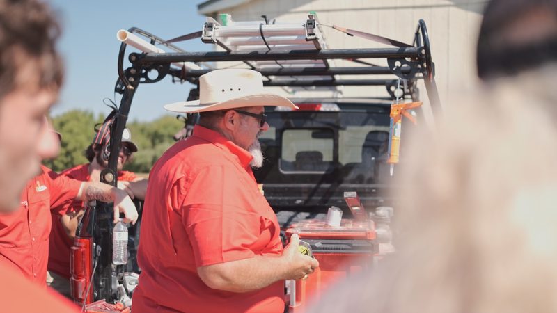 Tool cases and drink bottles on the open tailgate of a work truck, a Bell crew member in a straw hat alongside