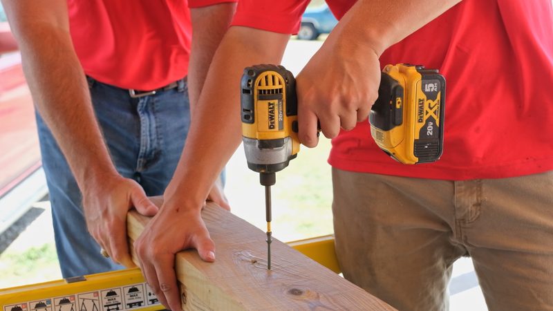 A cordless impact driver sinking a screw into a board while a second pair of hands holds the board steady