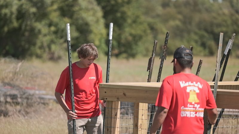 Two Bell crew members carrying steel line posts beside a new wood-framed wire fence, open pasture behind