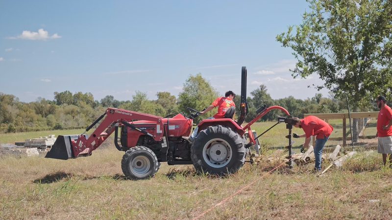 A compact tractor with a post-hole auger on the three-point hitch working along a fence line, crew alongside