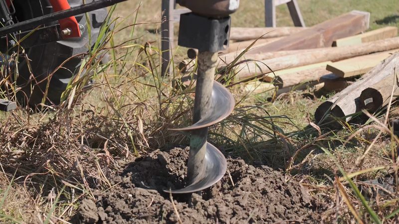 Close-up of a post-hole auger bit turning in fresh dirt beside stacked timber