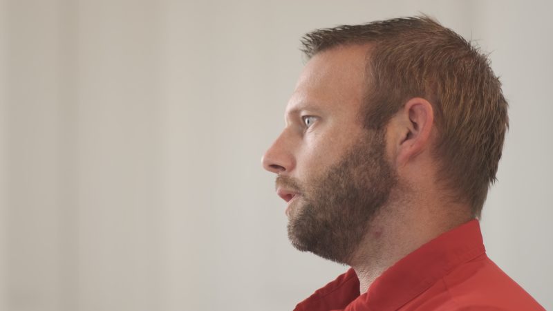 A member of the Bell team in close profile against a plain pale wall, mid-sentence.