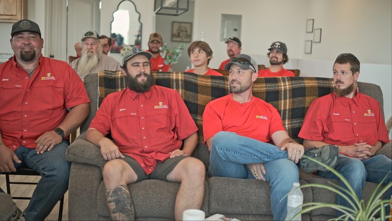 Bell crew in red company shirts seated on a couch and chairs indoors, listening, with others standing behind them