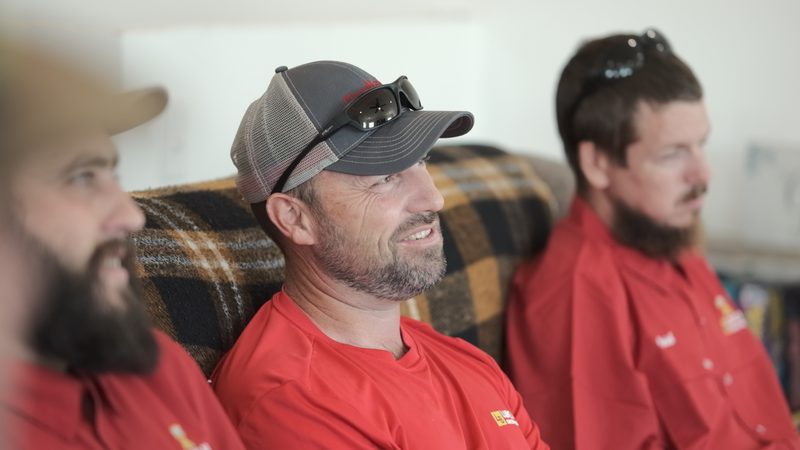 Three people in red company shirts seated indoors, one of them smiling, a cap with sunglasses pushed up on the brim.