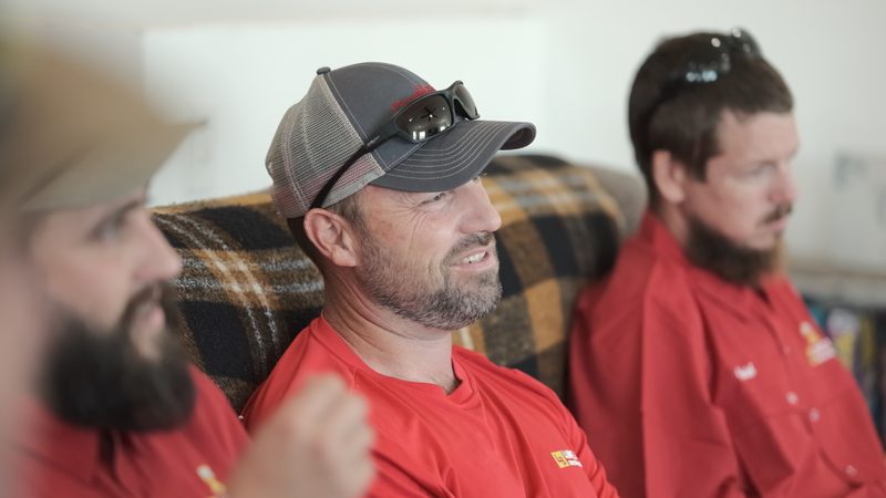 Close framing of Bell crew seated and listening, one in a mesh cap with sunglasses pushed up