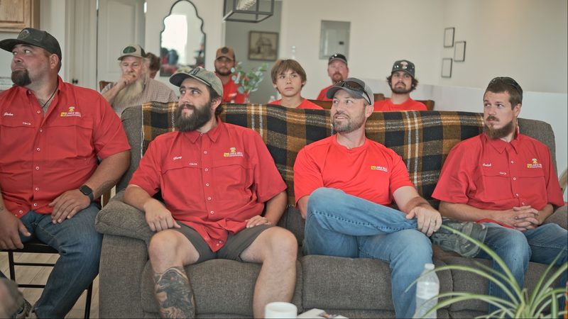 Crew in red company shirts seated on couches and chairs indoors, listening, with more people standing behind them.