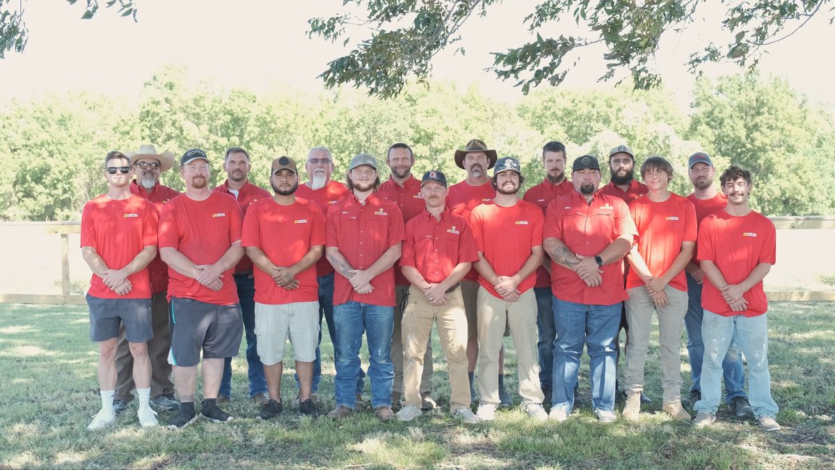Bell crew members in red company shirts lined up in two rows outdoors under a tree.