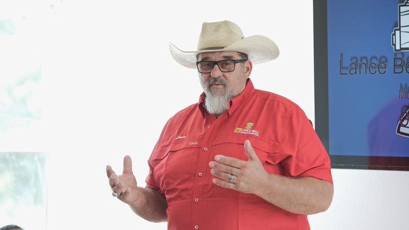 A man in a straw hat and red company shirt speaking to the room, a wall-mounted screen behind him.